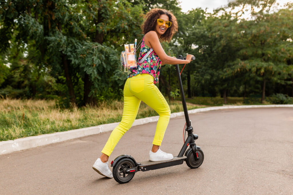 young stylish black woman having fun park riding electric kick scooter summer fashion style colorful hipster outfit wearing backpack yellow trousers sunglasses x